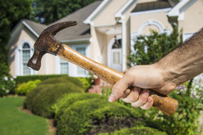 Mans hand holding hammer in front of a house indicating home improvement and maintenance.